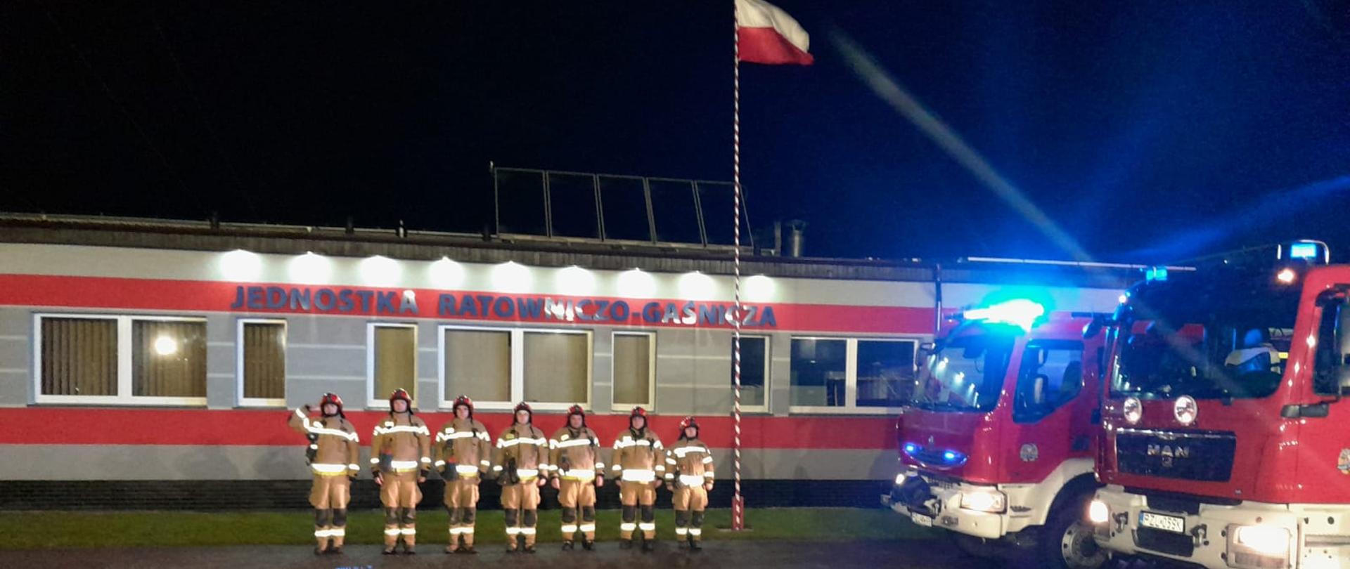 Na zdjęciu strażacy z JRG Złotów oddający hołd strażakom. W tle budynki JRG Złotów po prawej stronie samochody strażackie z włączonymi sygnałami.