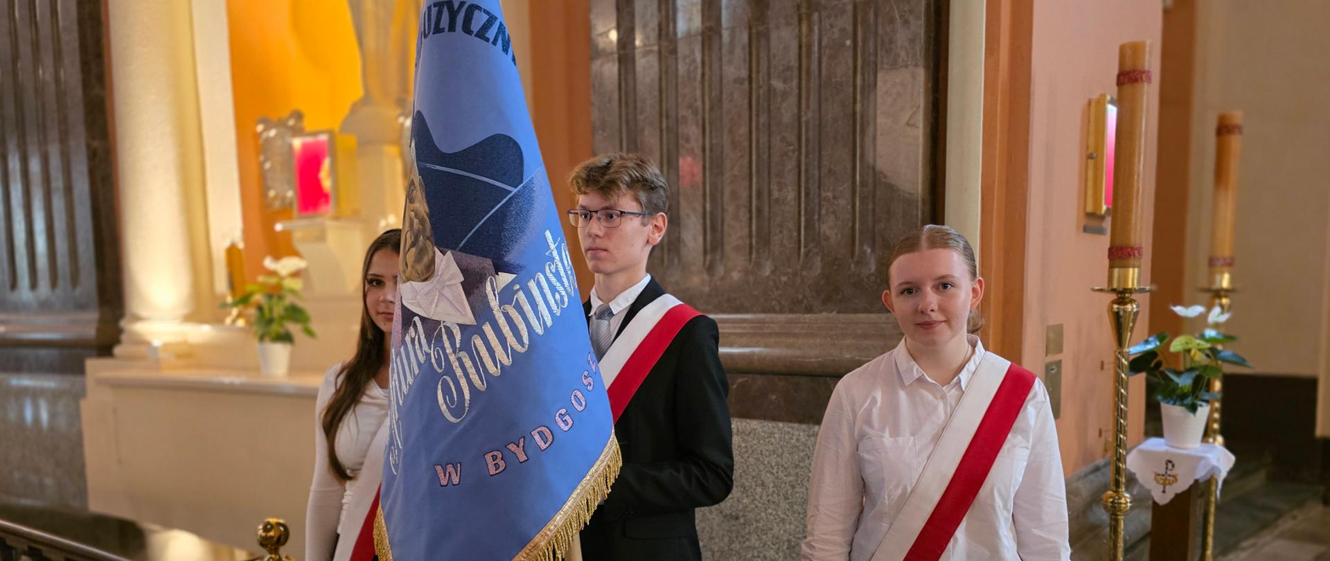 na zdjęciu dwie dziewczyny w białych koszulach i czarnych spodniach pośrodku chłopiec w okularach o blond włosach trzymający w rękach sztandar szkoły w kolorze niebieskim obszyty zlotymi frędzlami