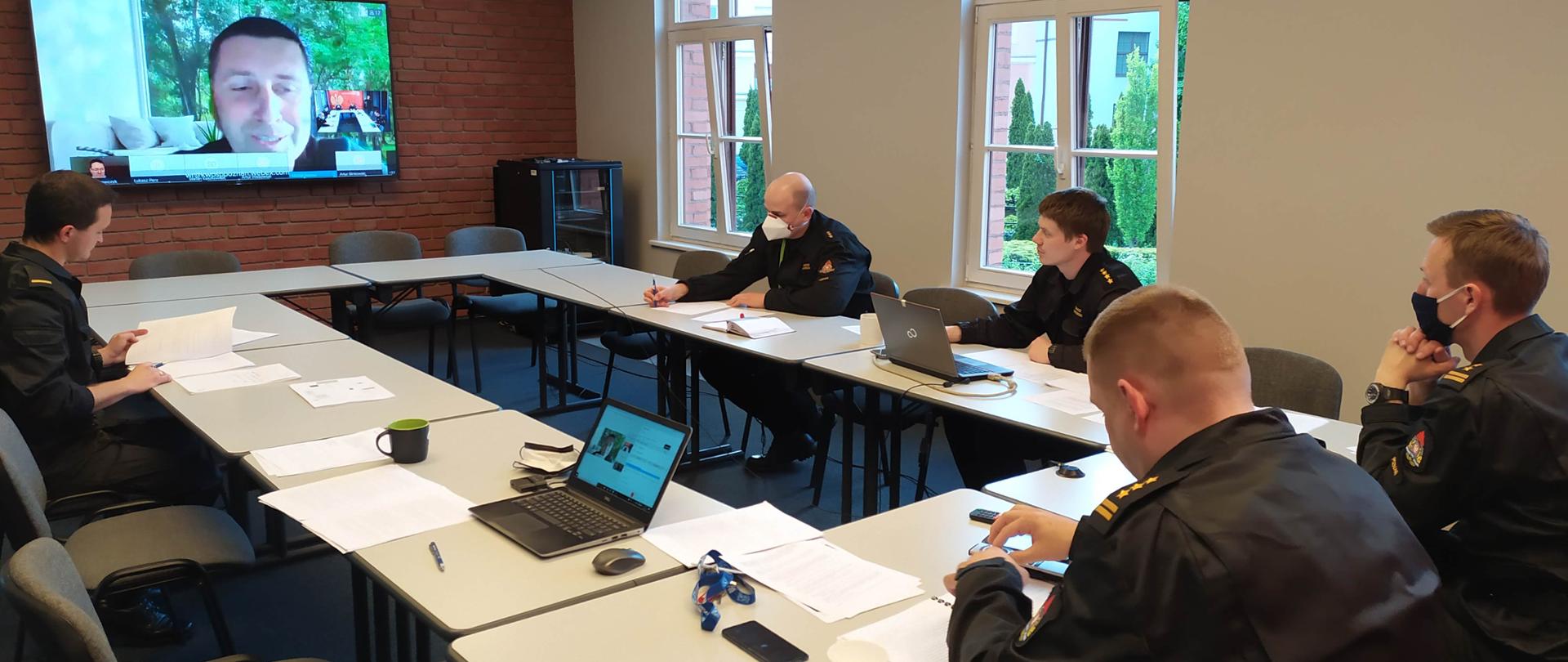 strażacy siedzący w czarnych mundurach przed dużym telewizorem,