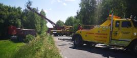 Na zdjęciu widać dwa holowniki drogowe, które wyciągają samochód ciężarowy po wypadku z przydrożnego pola. Ciężarówka zatrzymała się przed drzewami i stoi na polu. Jest słoneczny dzień