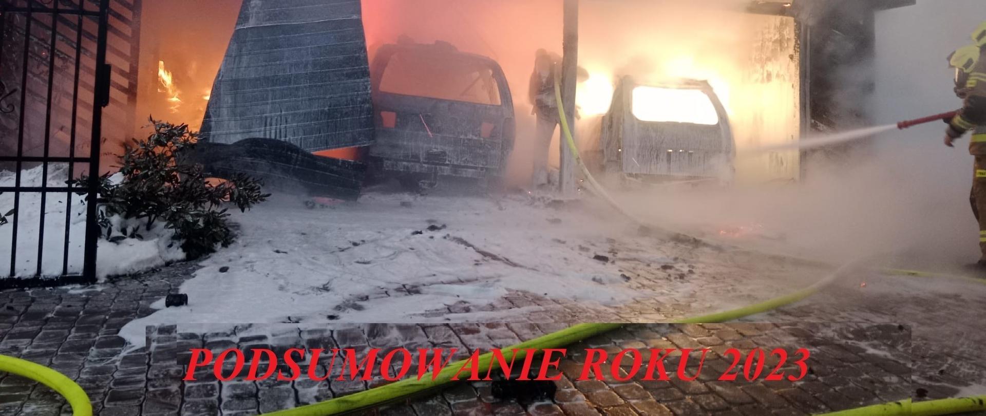 Fotografia wykonana w pochmurny dzień podczas gaszenia wiaty garażowej i samochodów w środku. Strażacy ubrani w ubrania specjalne podają pianę gaśniczą. Na dole zdjęcia napis "Podsumowanie roku 2023"
