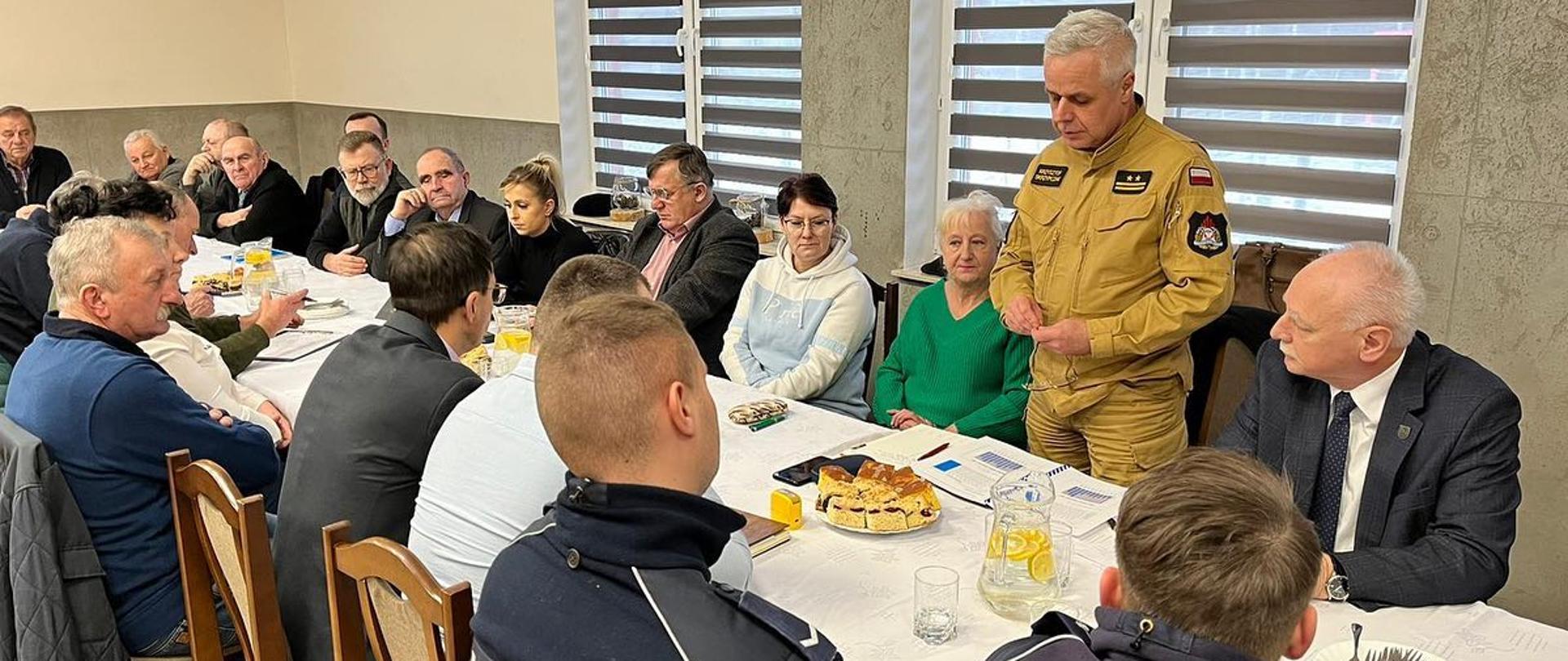 Spotkanie z sołtysami Na sali wiejskiej, po obu stronach stołu siedzą kobiety i mężczyźni - sołtysi, przedstawiciele władz gminy oraz dwóch policjantów. Na tle okna częściowo przysłoniętego roletami, stoi strażak w ubraniu służbowym w kolorze piaskowym, który przedstawia zebranym informacje z zakresu ochrony przeciwpożarowej. Na stole nakrytym białym obrusem stją dzbanki z napojami i talerze z ciastem .
