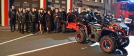 Kolorowa fotografia wykonana na zewnątrz wieczorem. Przedstawia strażaków OSP w mundurach oraz władze gminy i powiatu oraz quad na placu przy bramach garażowych jednostki OSP Jasienica Rosielna.