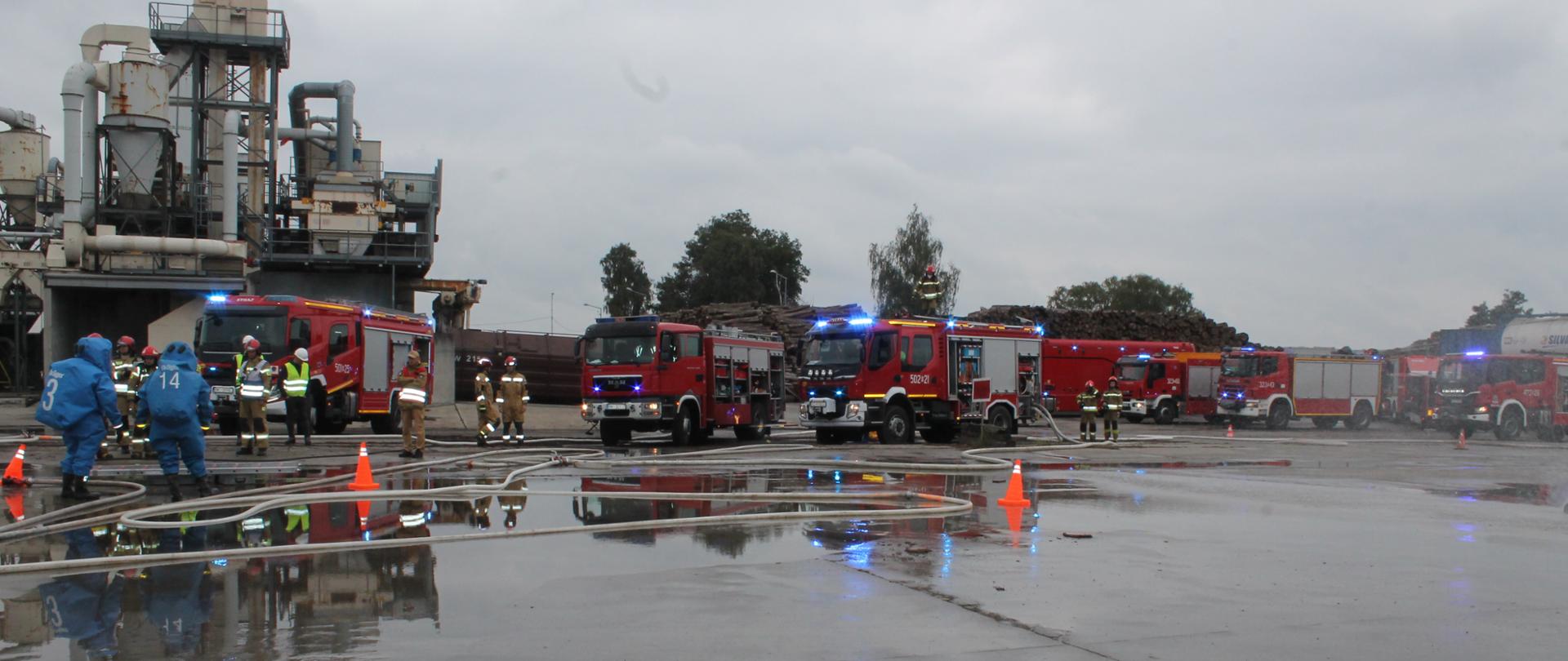 Szeroki plan ćwiczeń na zalanym placu przed instalacjami przemysłowymi. W deszczowym, pochmurnym otoczeniu widać szereg czerwonych wozów strażackich ustawionych w rzędzie, z włączonymi niebieskimi światłami sygnalizacyjnymi. Na mokrej, odbijającej światło nawierzchni widoczne są węże gaśnicze i pomarańczowe pachołki drogowe. Po lewej stronie widać strażaków w niebieskich kombinezonach chemicznych i żółto-brązowych strojach bojowych.