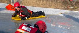 Na zdjęciu widoczny ratownik w przeręblu. Drugi ratownik zbliża się do niego na żółtej desce. W tle wóz strażacki.