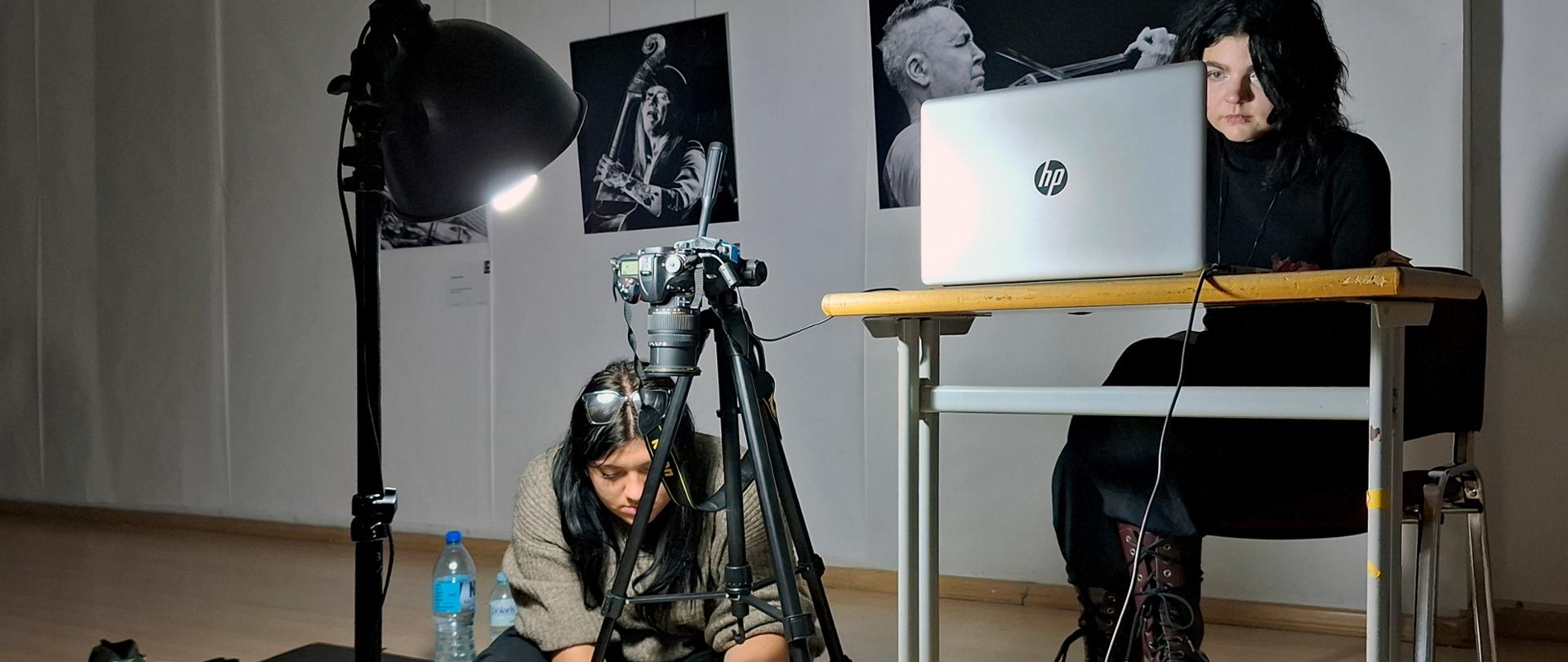 Dwie młode osoby. Jedna pracuje przy komputerze, druga siedzi przy aparacie fotograficznym, obok włączona lampa studyjna. Za nim na ścianie czarno-białe zdjęcia