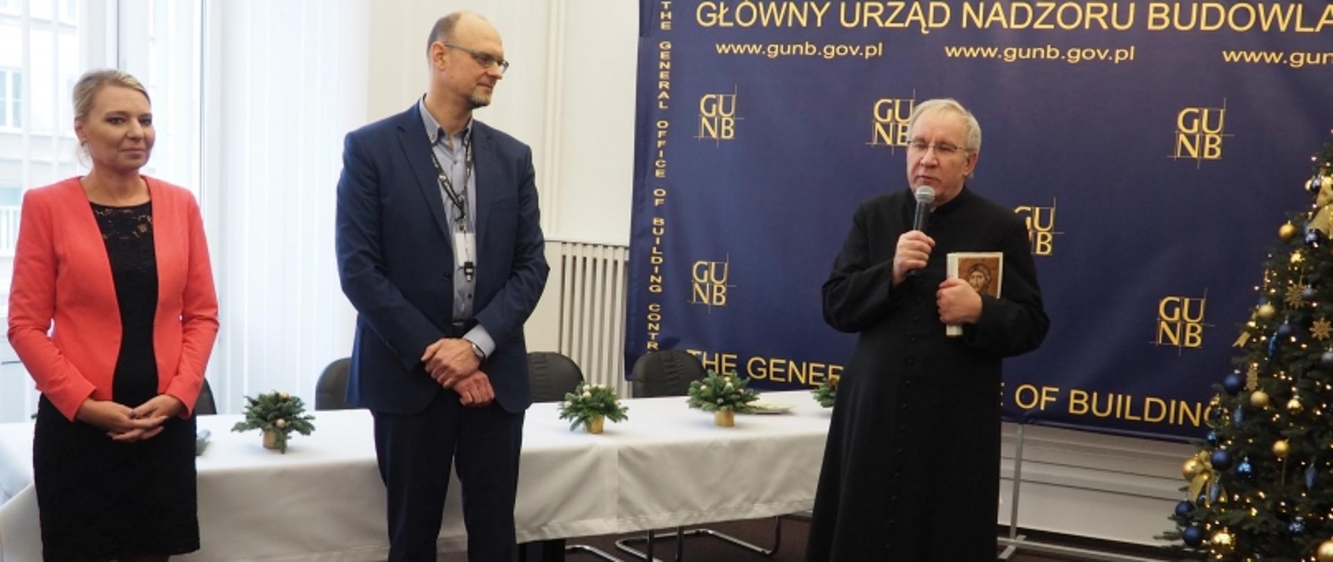 Trzy osoby przy stole z dekoracjami świątecznymi i choinką, w tle baner GUNB z nazwą urzędu i adresem www.gunb.gov.pl.