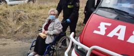 Starsza kobieta na wózku inwalidzkim transportowana przez strażaka. Z prawej strony fragment samochodu strażackiego. 