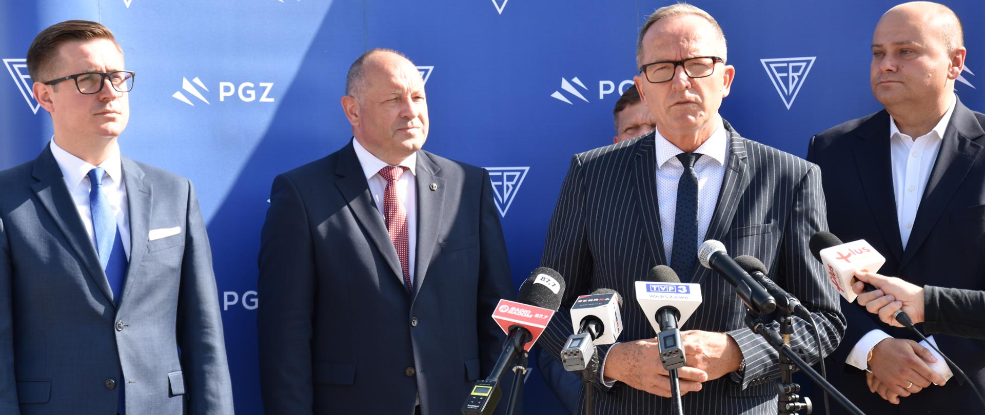 Na zdjęciu widać czterech elegancko ubranych mężczyzn. Jeden z nich stoi przy mikrofonach i przemawia podczas otwarcia wystawy plenerowej na placu Jagiellońskim w Radomiu upamiętniającej 95 lat działalności Fabryki Broni w Radomiu