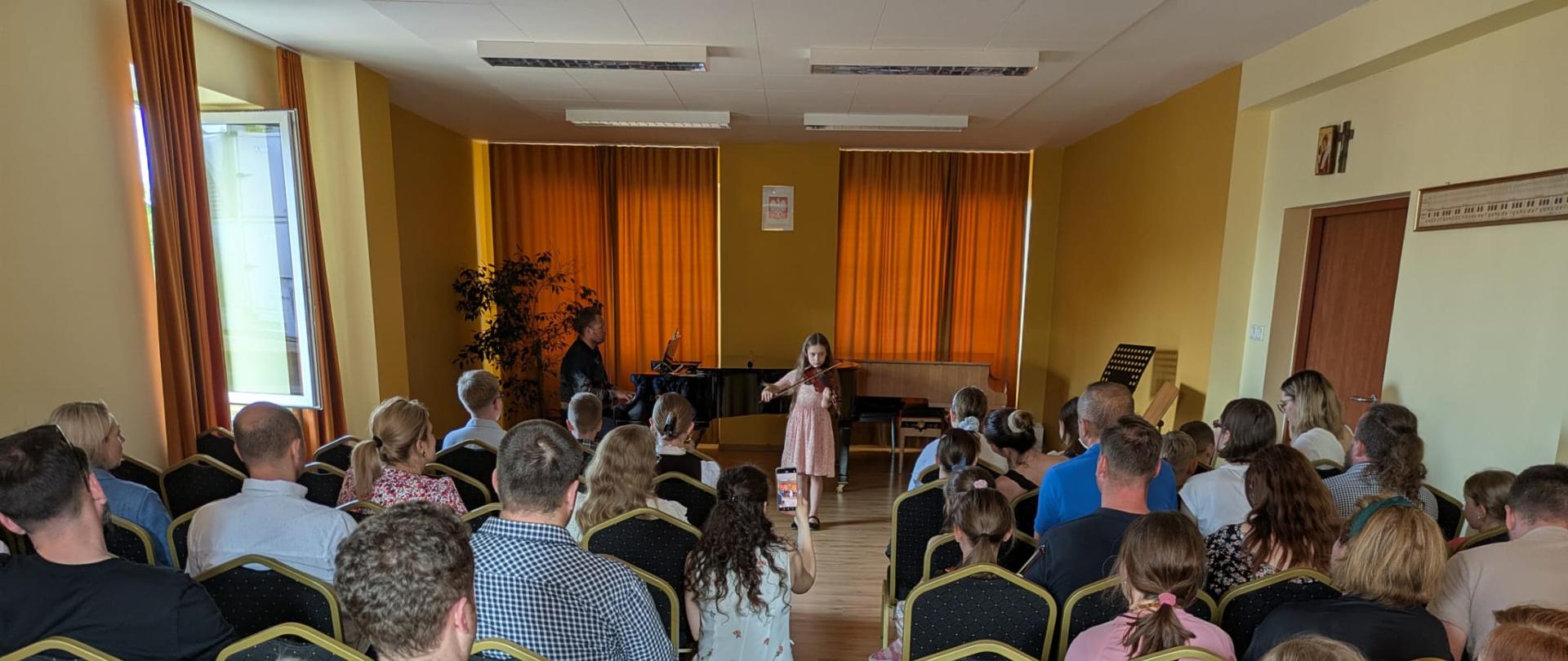 Aula podczas koncertu zapełniona publicznością. Na scenie gra skrzypaczka, której akompaniuje na fortepianie nauczyciel .