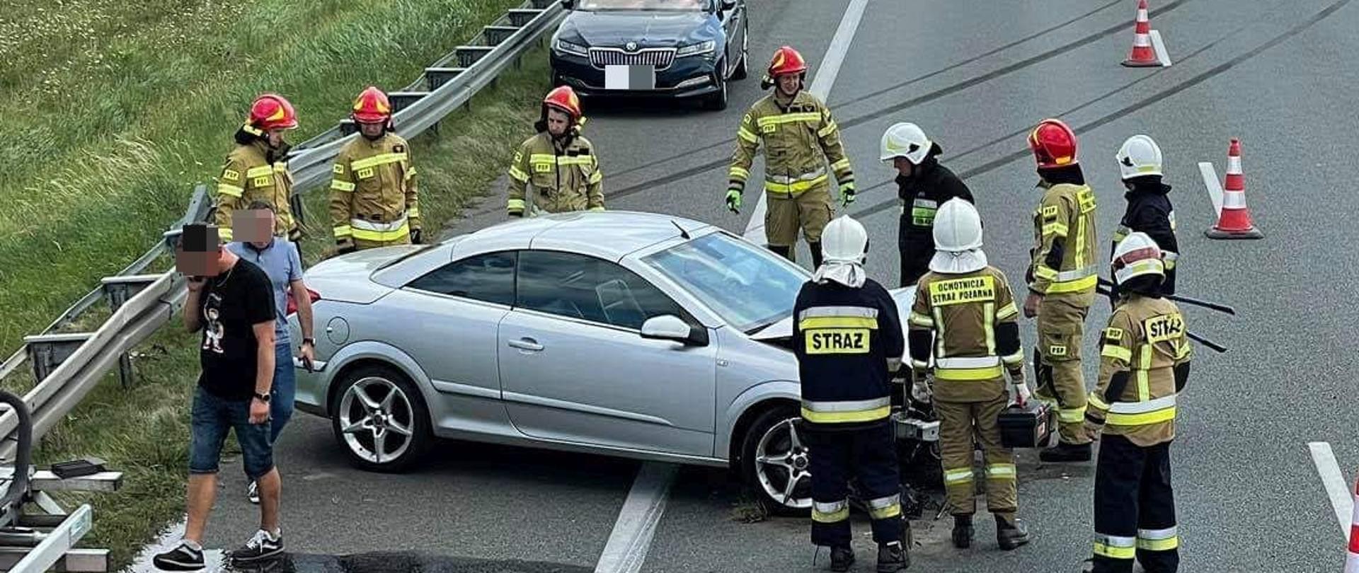 Na autostradzie a-2 znajduje się uszkodzony srebrny samochód, który stoi na pasie awaryjnym wokół niego stoi 10 strażaków z boku dwie osoby postronne jedna rozmawia przez telefon za srebrnym samochodem stoi inny zielony samochód oraz żółty pojazd służby autostradowej. Po pasie szybkim jedzie biały samochód, w oddali widać drzewa.