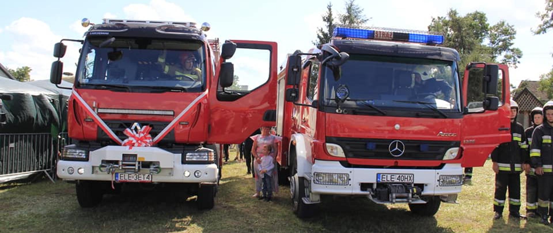 Na zdjęciu widać dwa czerwone samochody strażackie, które stoją na trawie. Obydwa samochody mają otwarte drzwi od strony kierowcy. Samochód z lewej strony zdjęcia ma z przodu przewiązaną biało-czerwoną kokardę. W samochodach siedzą kierowcy. Obok pojazdów stoją strażacy oraz dzieci. W tle widać namioty i drzewa. Jest widno.