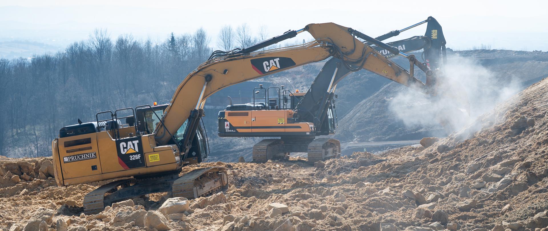 Na zdjęciu są dwie duże koparki gąsienicowe pracujące na skalistym stoku. Jedna z nich kruszy skały, unosząc chmurę pyłu. Teren jest jasny, kamienisty, a w tle widać nagie drzewa i lekko zamglony horyzont.