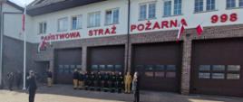 
Zdjęcie przedstawia uroczystość przed budynkiem Państwowej Straży Pożarnej w Ostrowi Mazowieckiej, w słoneczny dzień, prawdopodobnie podczas Dnia Flagi RP. Na pierwszym planie, na utwardzonym placu, stoi dwóch strażaków w galowych mundurach, salutujących. Jeden z nich znajduje się po lewej stronie kadru, zwrócony tyłem do widza, natomiast drugi, po prawej stronie, jest ustawiony profilem.
Za nimi, na środku kadru, stoi grupa strażaków w ciemnych, bojowych mundurach z żółtymi elementami. Budynek straży pożarnej jest biały z elementami ceglanymi i brązowym dachem. Nad trzema dużymi, brązowymi bramami garażowymi widnieje czerwony napis "PAŃSTWOWA STRAŻ POŻARNA" oraz numer alarmowy "998". Na fasadzie budynku, nad jedną z bram, wisi mała polska flaga. Z lewej strony budynku widoczny jest maszt flagowy, na którym również powiewa polska flaga. Na niebie widać kilka chmur.