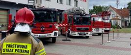 Z lewej strony widoczny jest strażak PSP, w tle widoczne są pojazdy specjalne PSP oraz Komenda Powiatowa. 