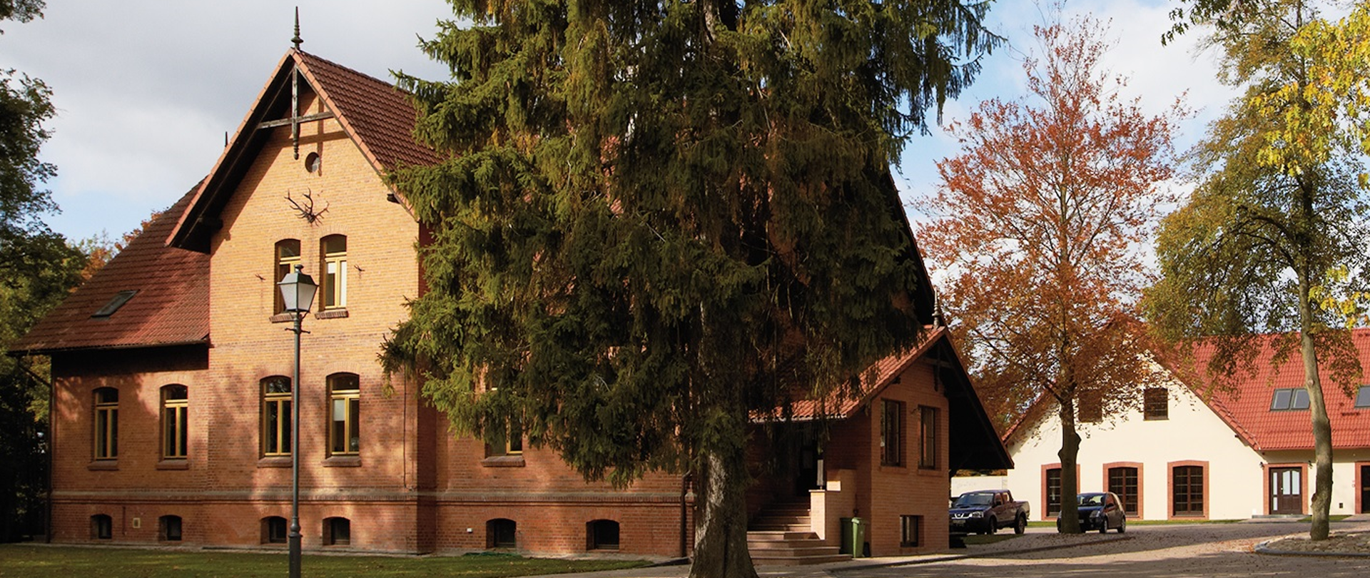 Ceglany budynek dwukondygnacyjny z końca XIX wieku na tle błękitnego nieba. Przed budynkiem od frontu rozpościera się zielony trawnik i latarnia, a od lewej strony widoczny jest parking. Centralnym punktem na zdjęciu jest przewyższający dwukrotnie wysokość budynku okazały świerk. Na drugim planie budynek edukacyjny z beżową elewacją i dachem w kolorze cegły. 