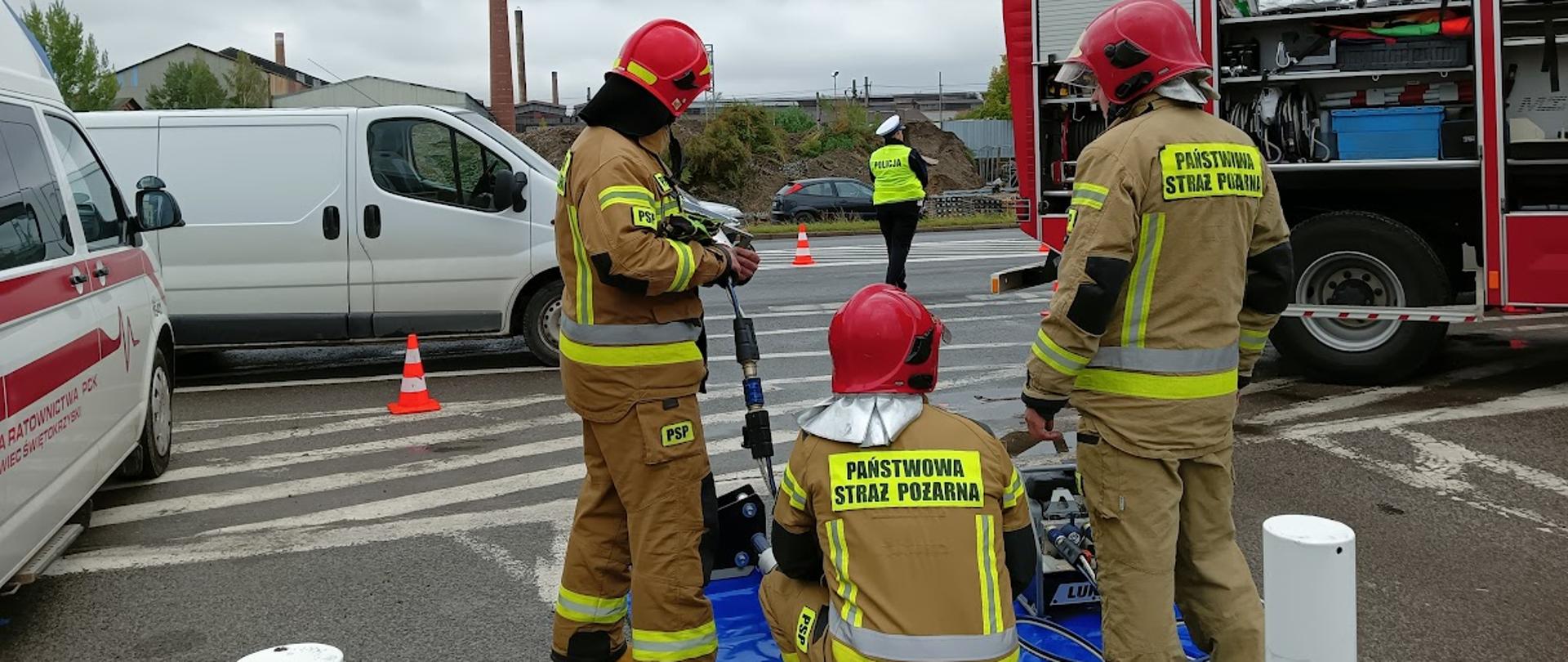 Strażacy przy stanowisku do pokazu sprzętu ratownictwa technicznego oraz patrol policji na drodze. 