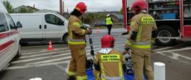 Strażacy przy stanowisku do pokazu sprzętu ratownictwa technicznego oraz patrol policji na drodze. 