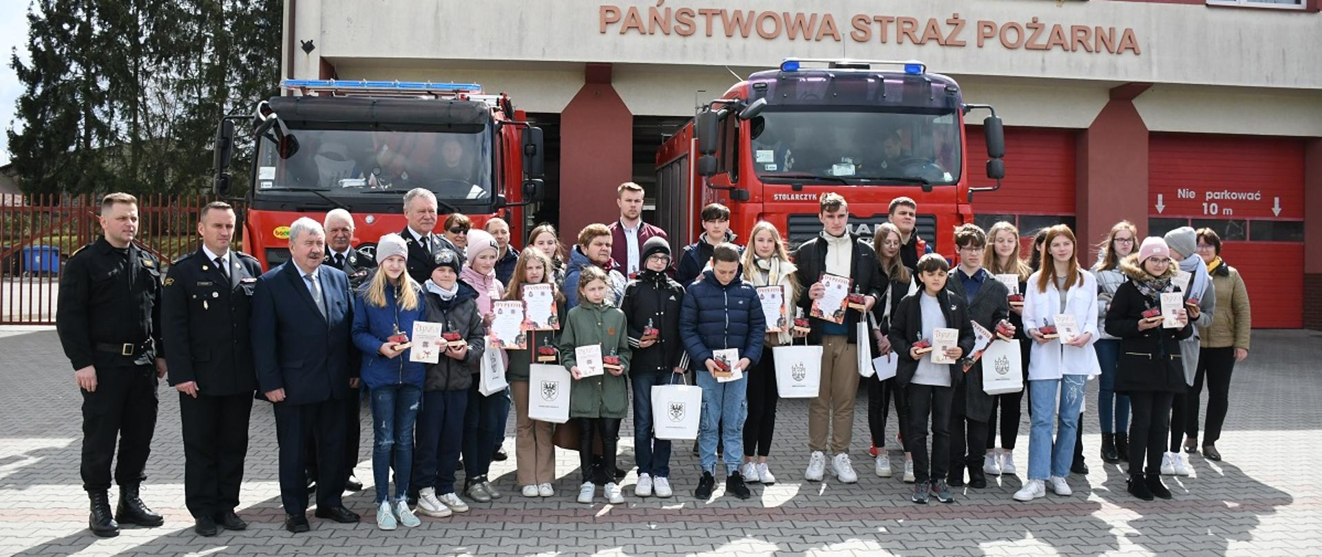 Zdjęcie uczestników turnieju na tle samochodów pożarniczych przed budynkiem JRG Miechów.