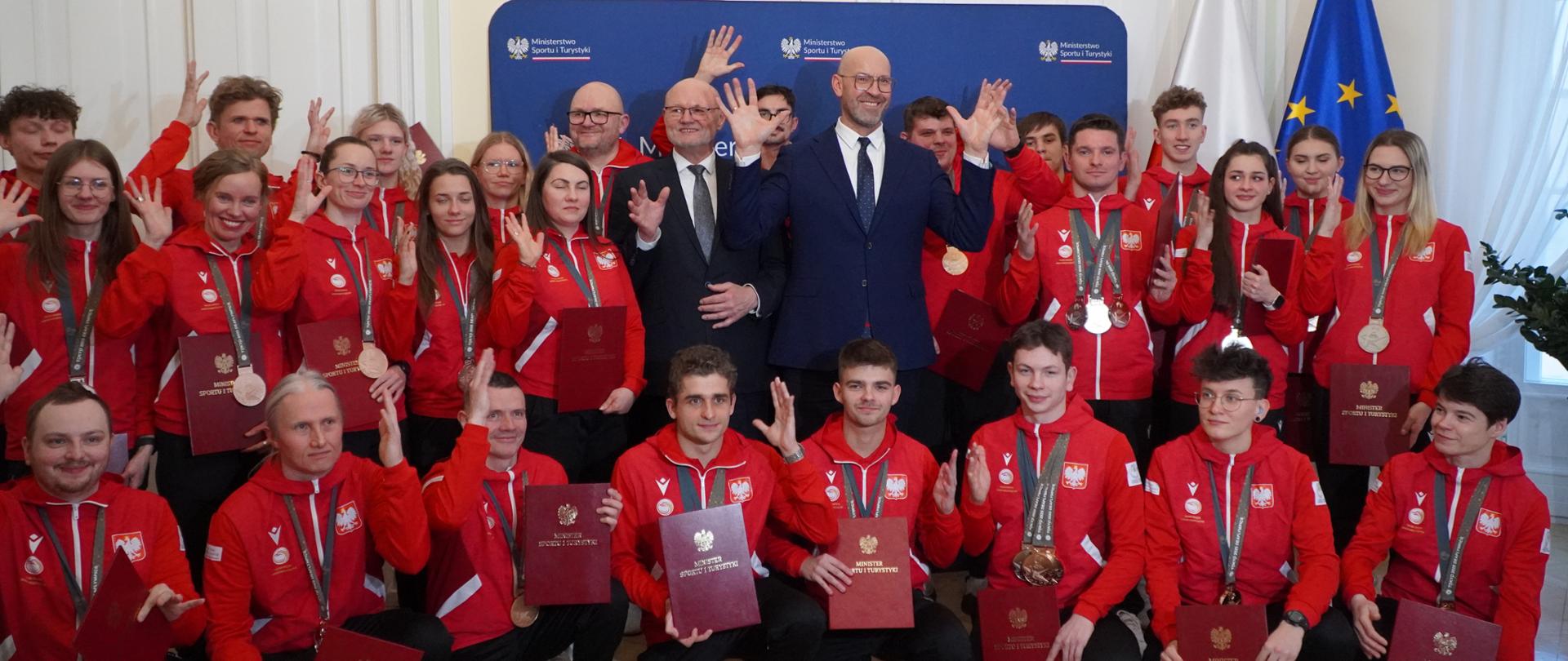 Zdjęcie zbiorowe medalistów XXV Letnich Igrzyskach Głuchych Tokio 2025 . Na środku stoją minister sportu i turystyki Jakub Rutnicki oraz podsekretarz stanu Ireneusz Nalazek. Za ich plecami widoczna ścianka MSiT oraz (z prawej strony zdjęcia) flagi Polski i UE.