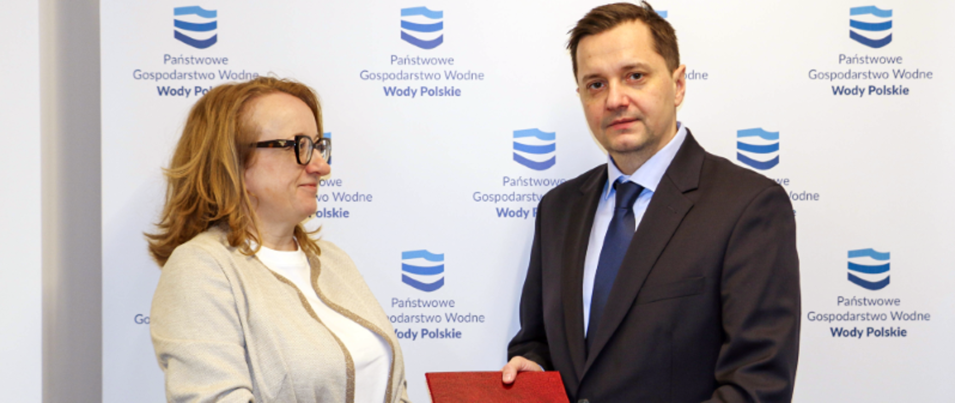 Prezes Wód Polskich Joanna Kopczyńska wręcza nominację na wiceprezesa pionu ochrony przed powodzią i suszą w Wodach Polskich Mateuszowi Balcerowiczowi. Stoją na tle ścianki z logiem Wód Polskich. 