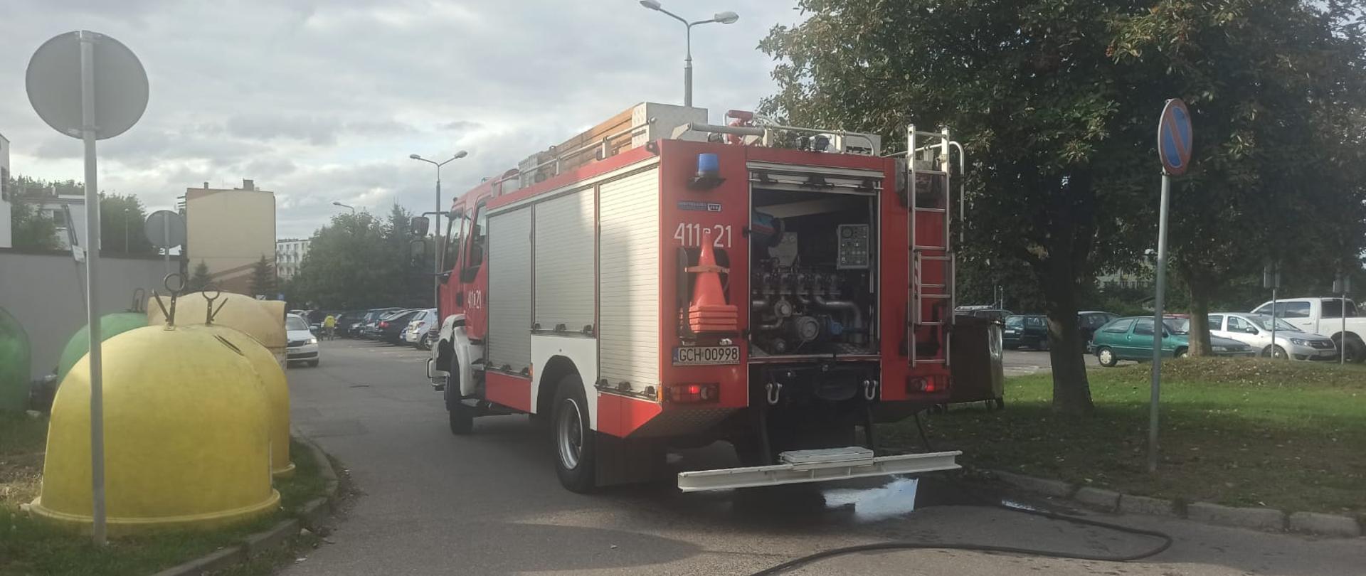 Czerwony samochód straży pożarnej stojący na drodze, otwarta tylna skrytka, w której znajduje się autopompa. Z lewej strony samochodu stoją żółte kontenery na śmieci, z prawej rośnie drzewo, w oddali widać bloki mieszkalne koloru brązowego. 