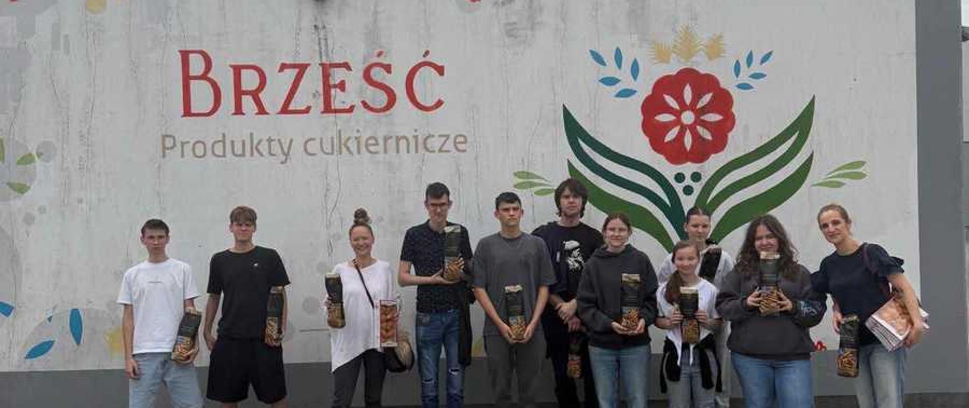 Zdjęcie przedstawia grupę dziesięciu osób stojących w jednym rzędzie na tle jasnej ściany budynku.Tło i otoczenieBudynek: Osoby stoją przed nowoczesnym, parterowym budynkiem o szarym wykończeniu dachu.Napis i logo: Na białej ścianie widnieje duży, czerwony napis „BRZEŚĆ” oraz mniejszy, szary pod nim: „Produkty cukiernicze”. Po prawej stronie napisu znajduje się duże, kolorowe logo przypominające tradycyjny, ludowy wzór z czerwonym kwiatem i zielonymi liśćmi.Podłoże: Grupa stoi na nawierzchni ułożonej z szarej kostki brukowej. Niebo nad nimi jest zachmurzone.Opis osóbSkład grupy: W rzędzie stoi pięciu młodych mężczyzn oraz pięć kobiet (w różnym wieku, od nastolatek po dorosłe osoby).Ubiór: Wszyscy są ubrani w luźne, codzienne stroje – t-shirty, bluzy z kapturem, jeansy, krótkie spodenki oraz buty sportowe.Paczki z produktami: Większość osób trzyma w dłoniach podłużne, pionowe opakowania (prawdopodobnie z produktami cukierniczymi firmy Brześć, takimi jak słomka ptysiowa).Ustawienie i pozy: Uczestnicy stoją blisko siebie, patrzą w stronę obiektywu i lekko się uśmiechają. Pozują w sposób naturalny i swobodny.