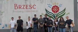 Zdjęcie przedstawia grupę dziesięciu osób stojących w jednym rzędzie na tle jasnej ściany budynku.Tło i otoczenieBudynek: Osoby stoją przed nowoczesnym, parterowym budynkiem o szarym wykończeniu dachu.Napis i logo: Na białej ścianie widnieje duży, czerwony napis „BRZEŚĆ” oraz mniejszy, szary pod nim: „Produkty cukiernicze”. Po prawej stronie napisu znajduje się duże, kolorowe logo przypominające tradycyjny, ludowy wzór z czerwonym kwiatem i zielonymi liśćmi.Podłoże: Grupa stoi na nawierzchni ułożonej z szarej kostki brukowej. Niebo nad nimi jest zachmurzone.Opis osóbSkład grupy: W rzędzie stoi pięciu młodych mężczyzn oraz pięć kobiet (w różnym wieku, od nastolatek po dorosłe osoby).Ubiór: Wszyscy są ubrani w luźne, codzienne stroje – t-shirty, bluzy z kapturem, jeansy, krótkie spodenki oraz buty sportowe.Paczki z produktami: Większość osób trzyma w dłoniach podłużne, pionowe opakowania (prawdopodobnie z produktami cukierniczymi firmy Brześć, takimi jak słomka ptysiowa).Ustawienie i pozy: Uczestnicy stoją blisko siebie, patrzą w stronę obiektywu i lekko się uśmiechają. Pozują w sposób naturalny i swobodny.