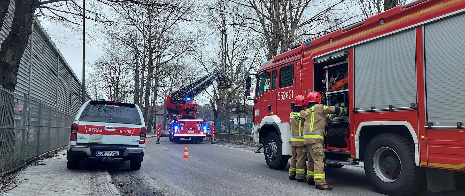 Dzień, nadłamane gałęzie drzewa zwisają niebezpiecznie nad drogą. W tle rozstawiona drabina mechaniczna z uniesionym koszem. Na pierwszym planie samochód strażacki, z którego skrytki dwóch strażaków wyjmuje sprzęt oraz samochód operacyjny.