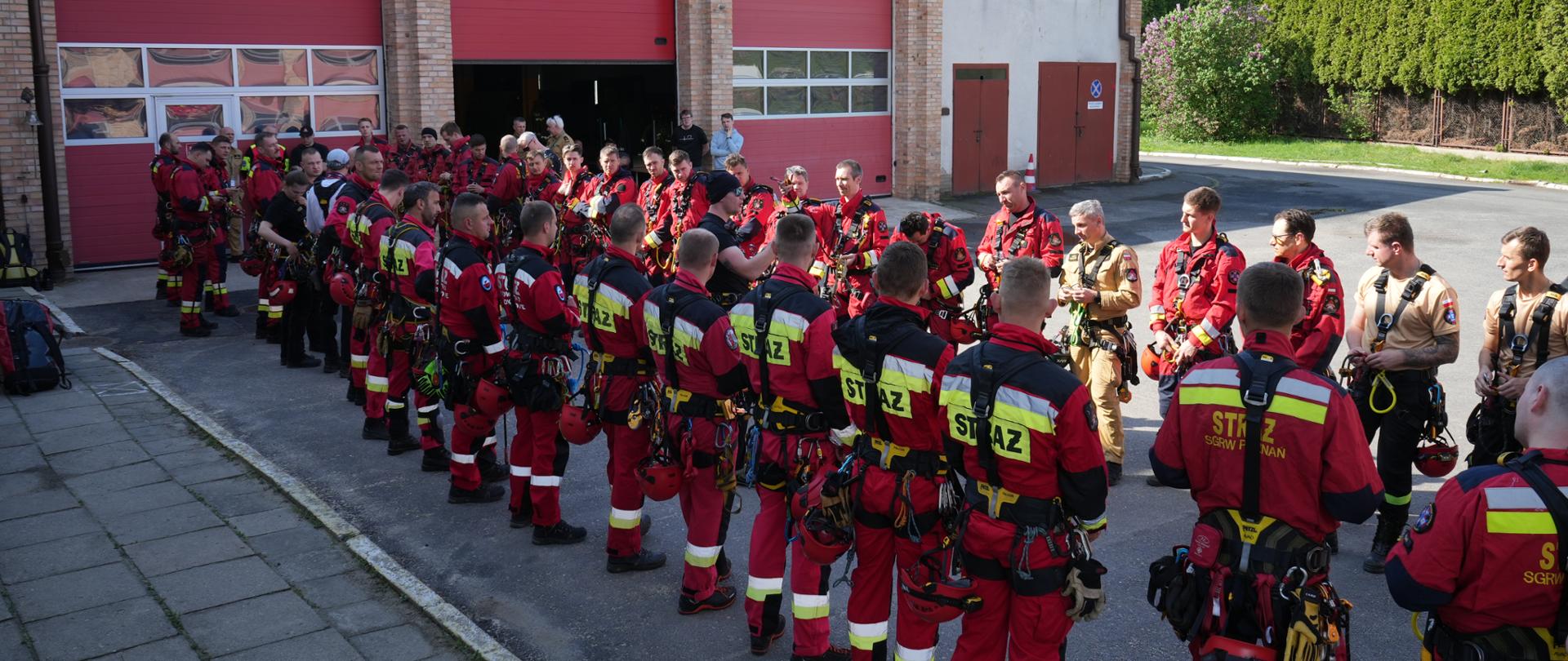 Liczna grupa ratowników SGRW w kombinezonach ćwiczeniowych zbiera się przed budynkiem remizy. Część osób analizuje sprzęt, trwa przygotowanie do ćwiczeń.
