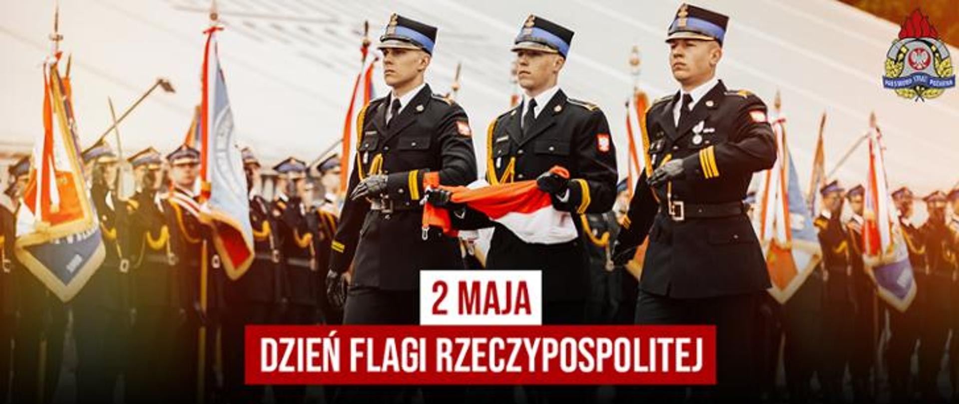 strażacy psp w umundurowaniu galowym maszerują w szyku niosąc flagę Polski. W tle widoczne sztandary i formacja honorowa. Na grafice napis: '' 2 maja- dzień flagi rzeczypospolitej"