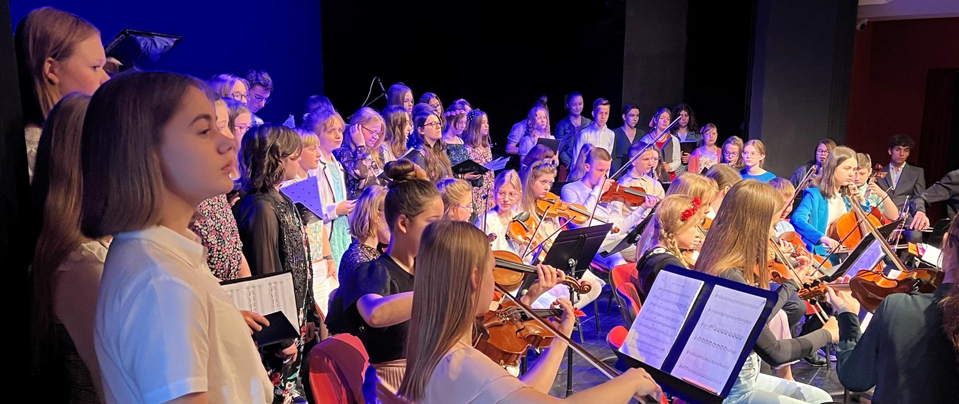 Kolorowe zdjęcie uczniów śpiewających w chórze oraz grających w orkiestrze podczas występu w Centrum Kultury Teatr w Grudziądzu