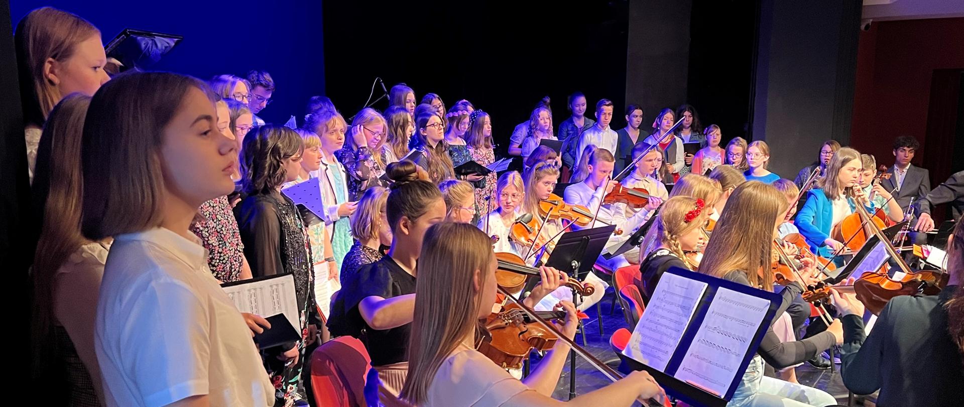 Kolorowe zdjęcie uczniów śpiewających w chórze oraz grających w orkiestrze podczas występu w Centrum Kultury Teatr w Grudziądzu