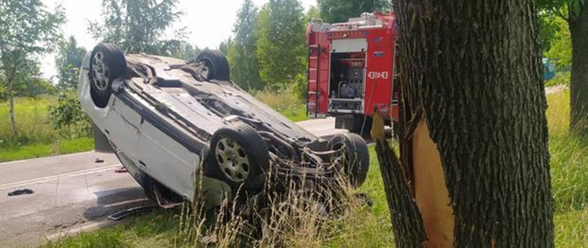 Widoczny samochód osobowy leżący na dachu po wypadku, obok zniszczone drzewo