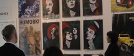 Na zdjęciu widoczne trzy osoby oglądające plakaty wiszące na ścianie.