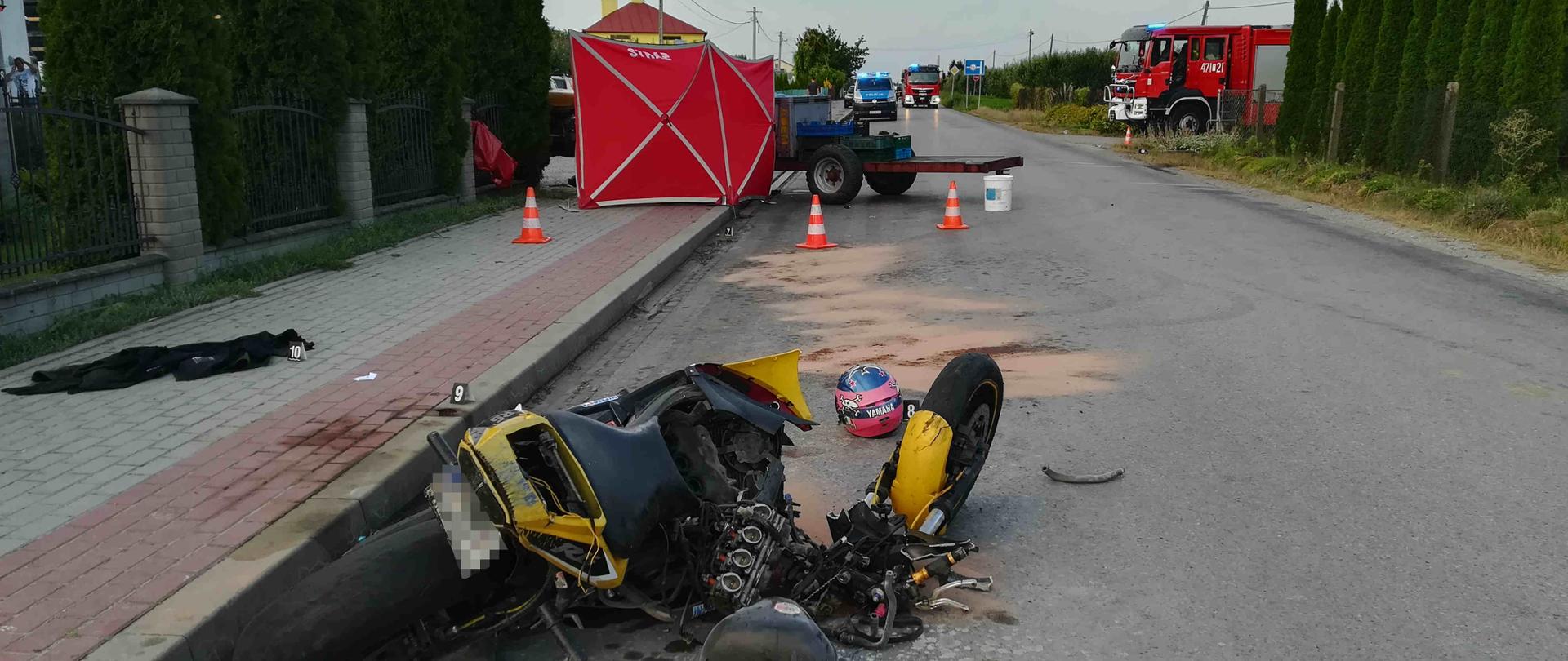 Zdjęcie zrobione w dzień. Na zdjęciu widać samochody strażackie i policyjny. W dalszej części zdjęcia widać rozbity motocykl, ciągnik rolniczy z przyczepką oraz parawan.
