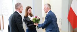 Na zdjęciu trzy osoby: minister Tomasz Siemoniak (po prawej), Oksana Poczobut (w środku) oraz Andrzej Poczobut (po lewej). Minister Tomasz Siemoniak wręcza Oksanie Poczobut bukiet kwiatów. Po prawej stronie zdjęcia widać flagę Polski (biało-czerwoną). Po lewej stronie zdjęcia widać szklane drzwi wejściowe z logo ABW.