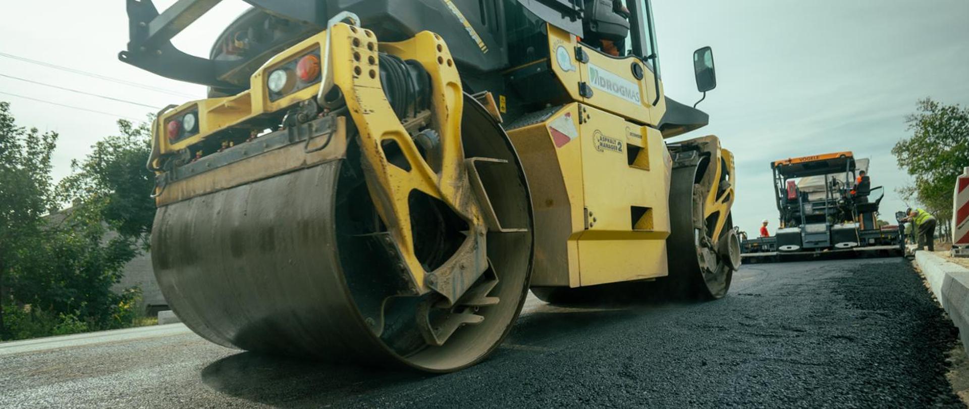 Walec drogowy zagęszcza świeżo ułożoną nawierzchnię asfaltową na budowanej drodze. W tle widoczna jest rozkładarka mas bitumicznych oraz pracownicy prowadzący roboty drogowe przy krawężniku.