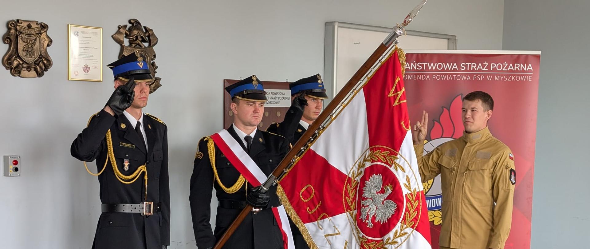 Na zdjęciu widoczna jest uroczysta przysięga strażacka. Po prawej stronie stoi strażak w mundurze koloru piaskowego, z uniesioną prawą ręką w geście ślubowania. Naprzeciw niego znajduje się poczet sztandarowy w mundurach galowych. Jeden strażak trzyma sztandar Państwowej Straży Pożarnej z godłem Polski i napisem „Bóg Honor Ojczyzna”, a dwóch oddaje honory salutując. W tle widać czerwony baner z napisem „Państwowa Straż Pożarna, Komenda Powiatowa PSP w Myszkowie.