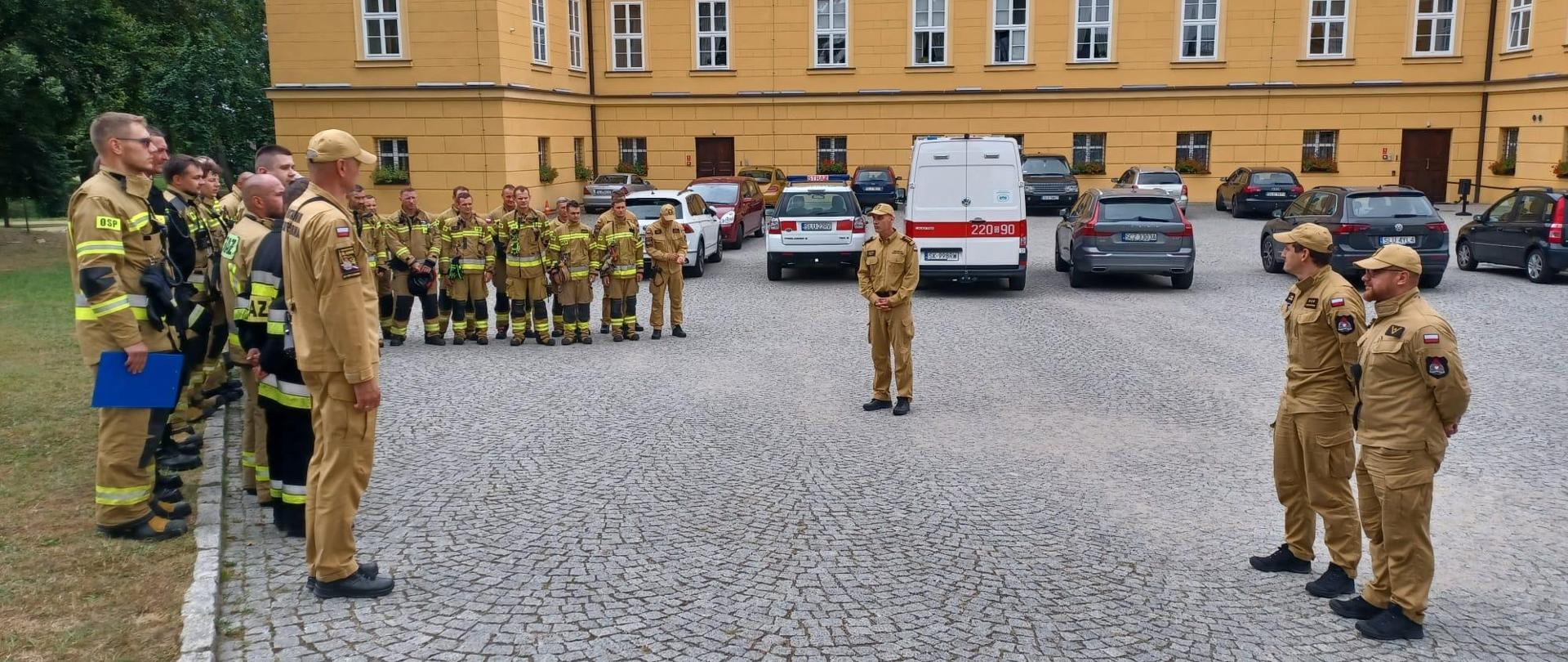 Zdjęcie przedstawia pododdział strażaków podczas odprawy, przed nimi w centrum zdjęcia wypowiada się Komendant Powiatowy PSP w Lublińcu, po prawej stronie dwóch strażaków w umundurowaniu służbowym z Komendy Wojewódzkiej PSP w Katowicach,w tle budynek kompleksu pałacowego oraz pojazdy cywilne oraz strażackie.