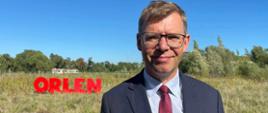 Wojewoda Artur Chojecki na tle pola na którym stoi wielki napis Orlen
