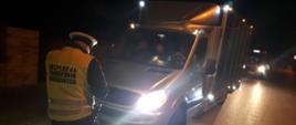 Na pierwszym planie: umundurowany funkcjonariusz mazowieckiej Inspekcji Transportu Drogowego stoi przed pojazdem zatrzymanym do kontroli. Inspektor trzyma w ręku i sprawdza dokumenty pobrane do kontroli od kierowcy. W tle: za pojazdem zatrzymanym do kontroli stoi oznakowany furgon mazowieckiej Inspekcji Transportu Drogowego.