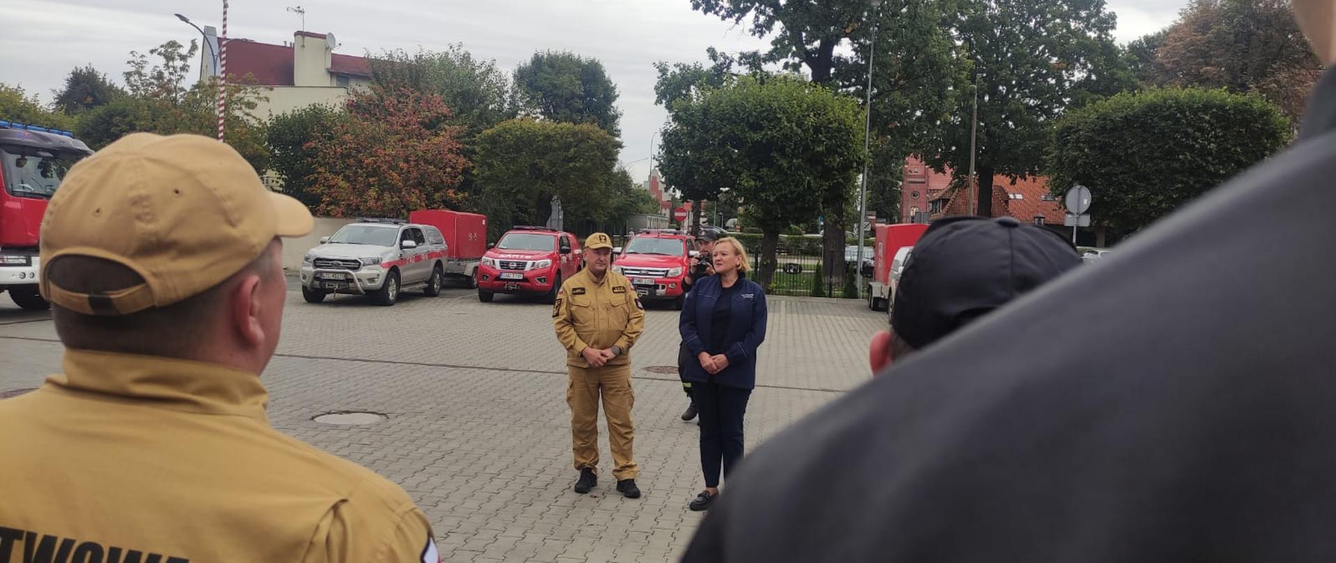 Pani Wicewojewoda oraz Pomorski Komendant Wojewódzki stoją naprzeciw strażaków, ujęcie zdjęcia z drugiego rzędu dwuszergu. W tle pojazdy strażackie.