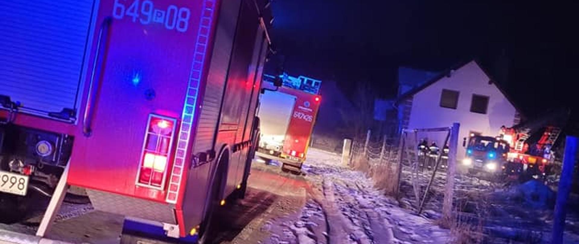 Noc, dwa samochody pożarnicze na drodze pokrytej śniegiem. W tle strażacy i podnośnik hydrauliczny.