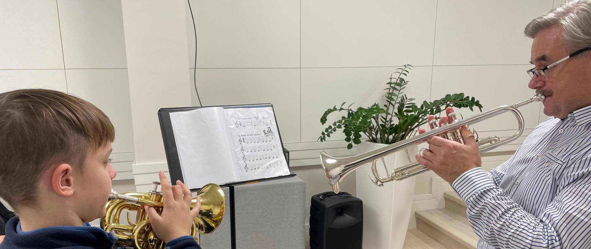 Zdjęcie przedstawia ucznia stojącego po lewej stronie i prowadzącego warsztaty trąbkowe – po prawej. Uczeń ubrany jest w ciemnoniebieską bluzę i gra na trąbce w kolorze złotym, natomiast prowadzący ma na sobie białą koszulę w ciemne paski i gra na trąbce w kolorze srebrnym. Pomiędzy nimi znajduje się pulpit z rozłożonymi nutami. Na białym tle widać stolik przykryty szarym pokrowcem, głośnik i białą donicę z zielonym kwiatem.