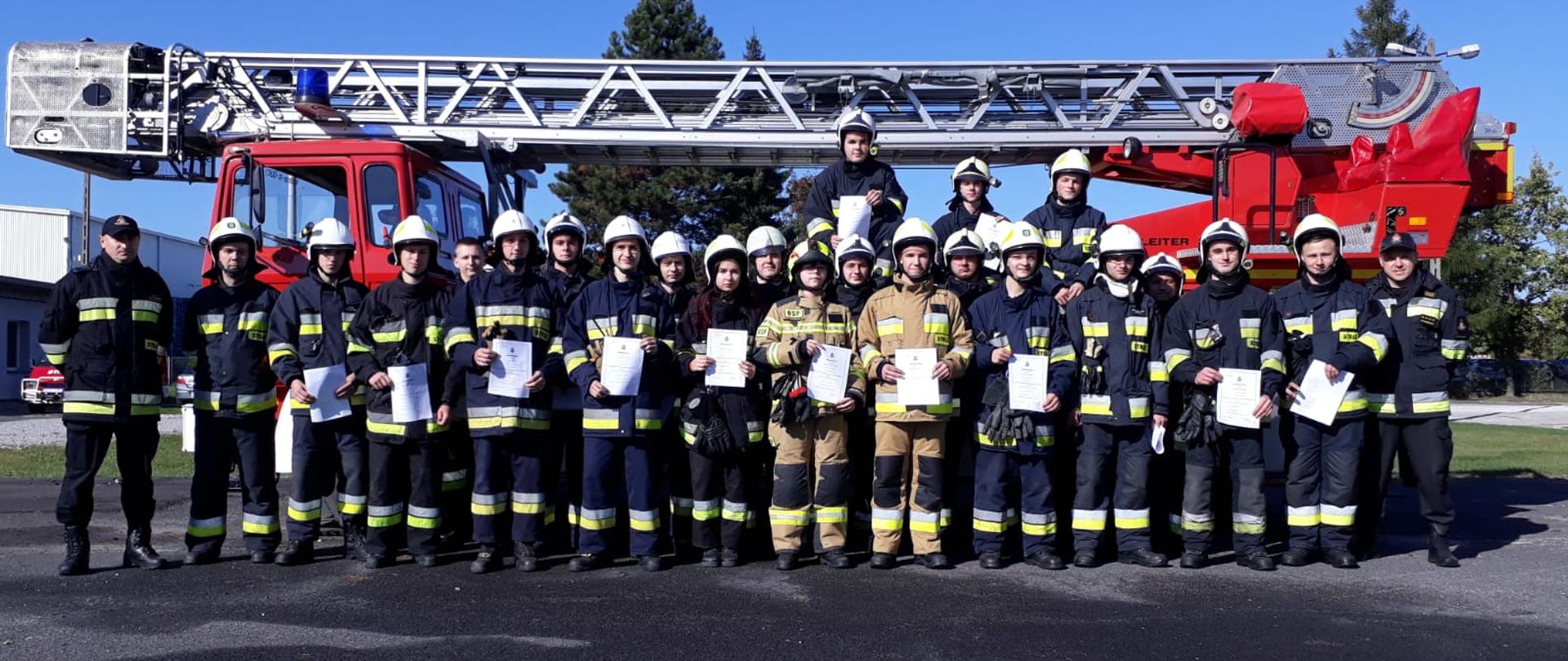 Na zdjęciu znajdują się strażacy w umundurowaniu specjalnym na tle drabiny pożarniczej