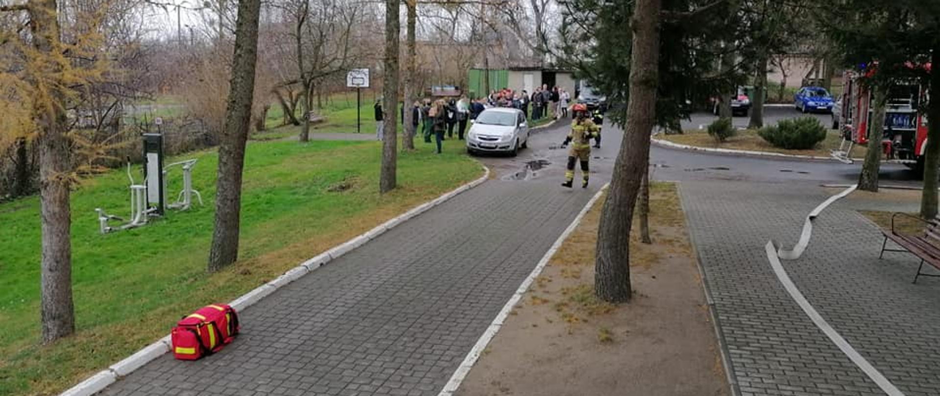 W tle widać grupę dzieci ustawionych przed budynkiem, strażaka w ubraniu specjalnym a po prawej stronie fragment wozu bojowego.
