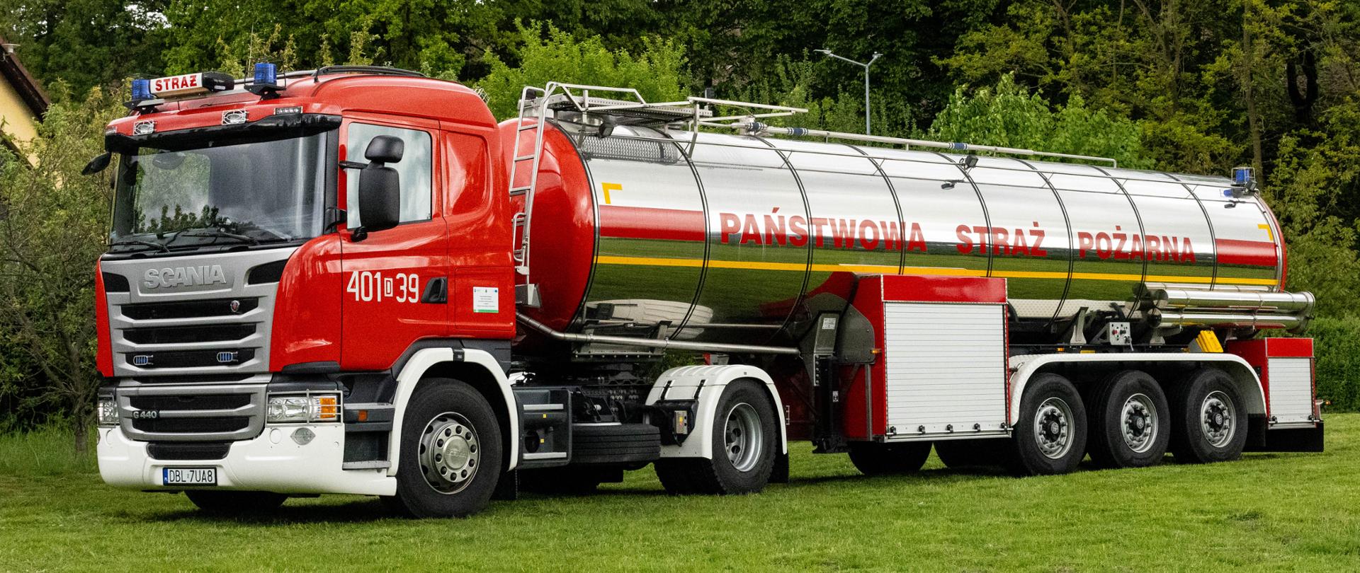 Ciężki ciągnik siodłowy Scania G440 (numer „401[D]39”) z podpiętą dużą, chromowaną naczepą-cysterną. Na błyszczącym boku cysterny widnieje czerwony napis „PAŃSTWOWA STRAŻ POŻARNA”. Cały zestaw stoi na zielonym terenie.