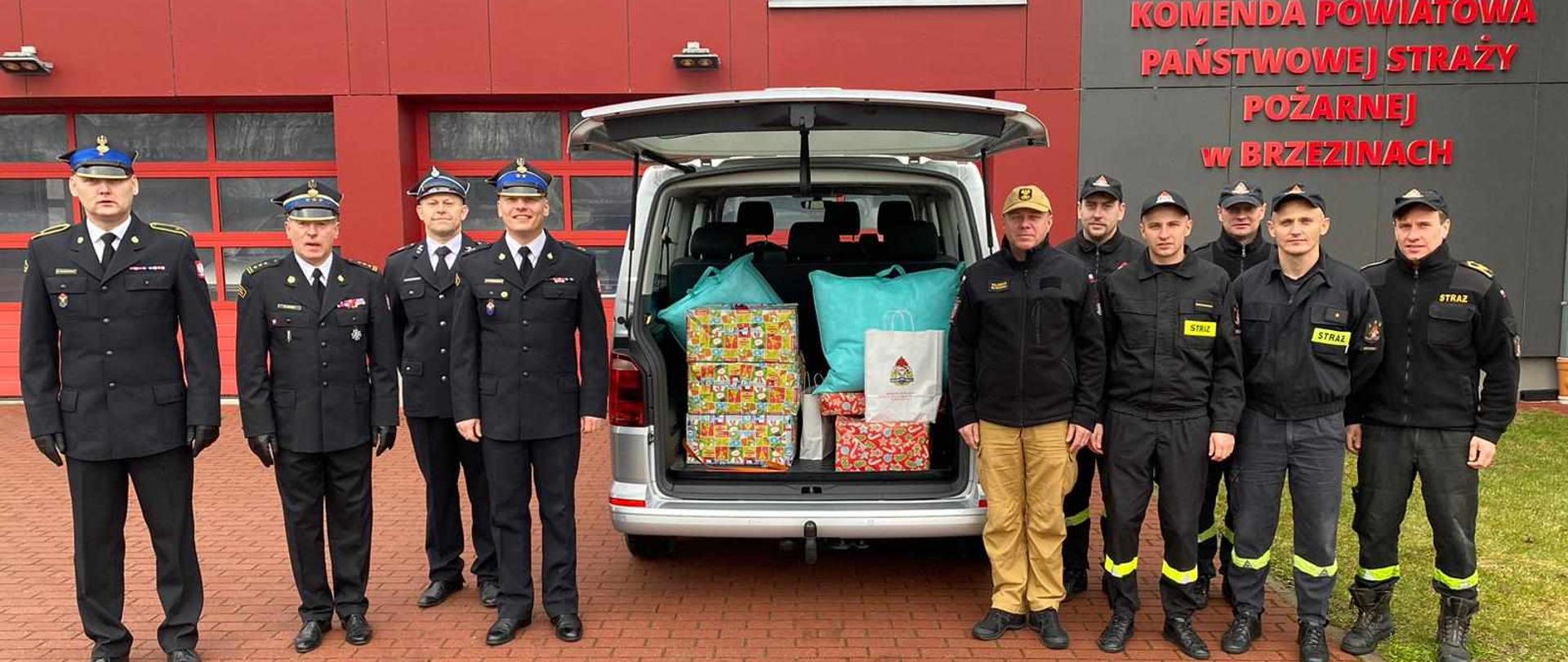 Plac przed komendą państwowej straży pożarnej w brzezinach w centrum zdjęcia znajduje się samochód w otwartym bagażnikiem gdzie w środku znajdują się dary dla rodziny ufundowane przez strażaków po obu stronach samochodu stoją strażacy z państwowej straży pożarnej w brzezinach