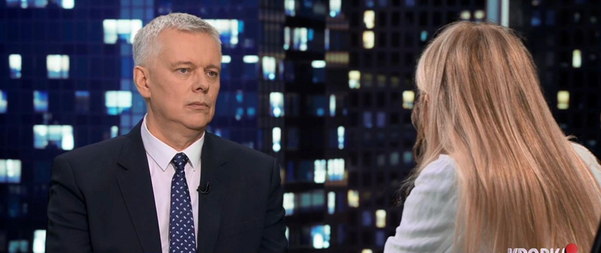 Kadr z programu telewizyjnego. Po lewej stronie siedzi minister Tomasz Siemoniak w ciemnym garniturze i krawacie, zwrócony w stronę rozmówczyni widocznej od tyłu po prawej stronie. W tle nocny widok na oświetlone okna budynków. Na dole ekranu znajdują się paski informacyjne z nagłówkiem dotyczącym wypowiedzi o akcji w Berlinie oraz logo stacji telewizyjnej.